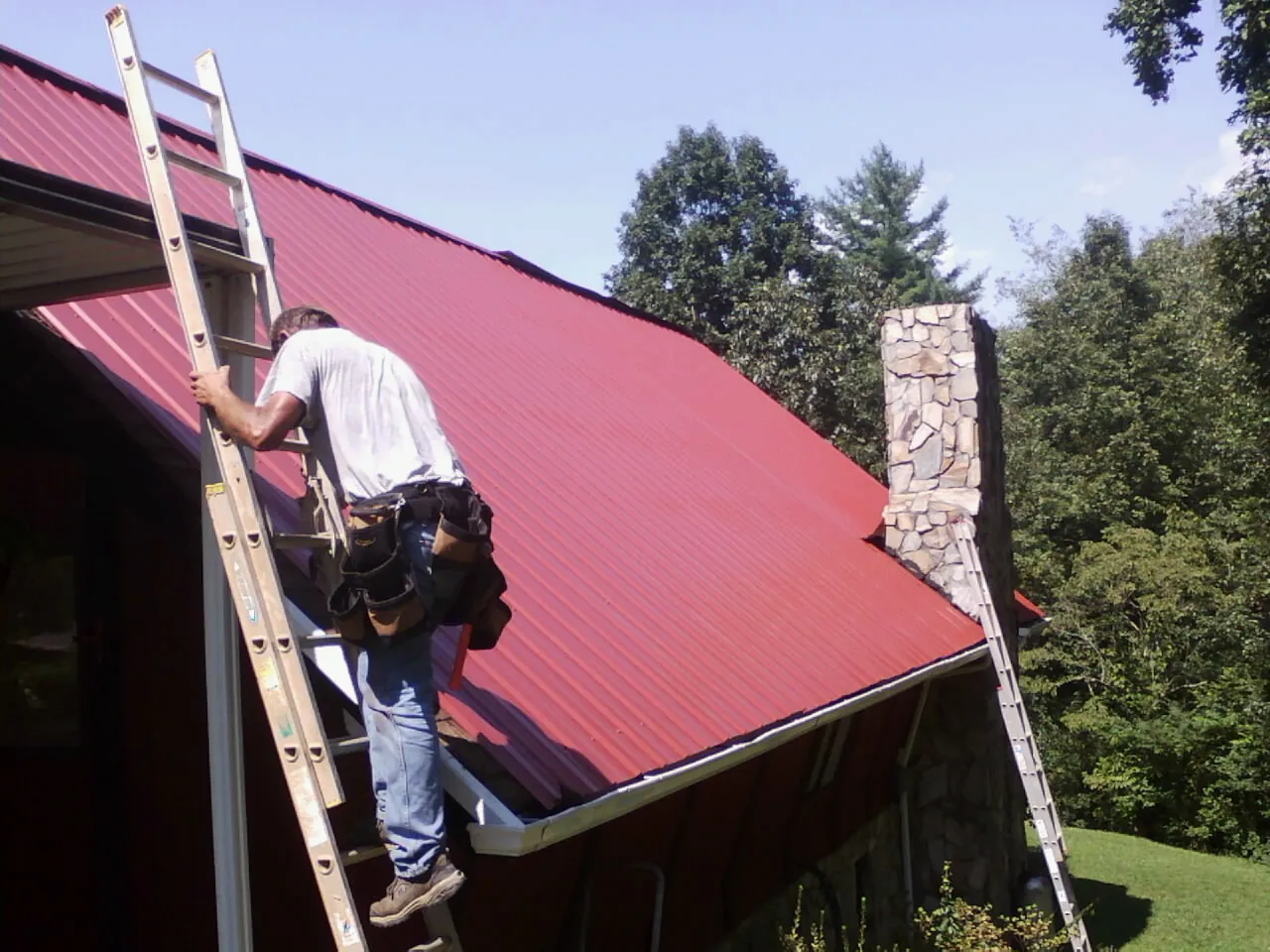 Roofer on a ladder beside a red metal roof and stone chimney — Soffit & Fascia Repair by Roofing Brothers in Mechanicsville